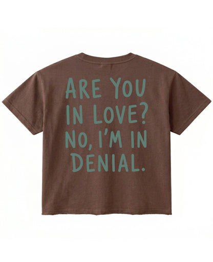 Back view of a brown boxy fit t-shirt with a raw unfinished bottom hem. It features large sage green text reading Are You In Love? No, I'm In Denial.