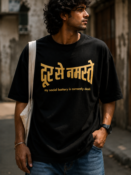 A man wearing a black t-shirt with gold Hindi text Dur Se Namaste and the subtitle 'my social battery is currently dead.' in English, styled with blue jeans and a white shoulder bag in an outdoor street setting.