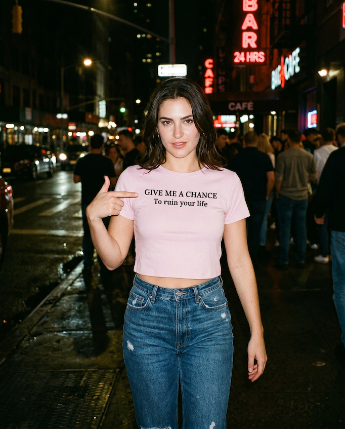 Woman wearing a pink Give Me A Chance To Ruin Your Life Baby Tee, t-shirt with text on a city street at night.