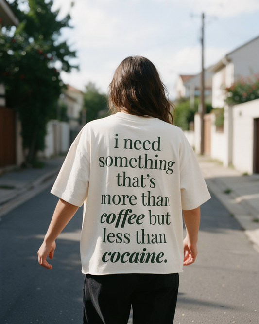 A person viewed from behind walking down a sunny residential street, wearing an oversized cream-colored t-shirt with dark green text that reads, 'i need something that's more than coffee but less than cocaine.'.