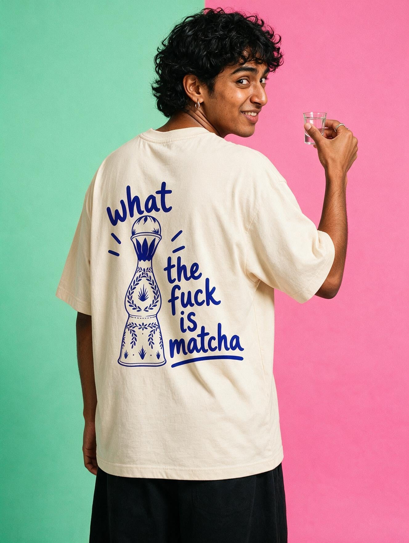 A man wearing an oversized cream-colored t-shirt with a blue graphic print on the back, posed against a split green and pink studio background.