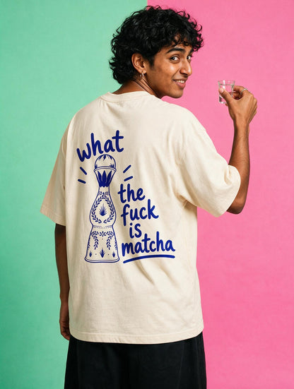 A man wearing an oversized cream-colored t-shirt with a blue graphic print on the back, posed against a split green and pink studio background.