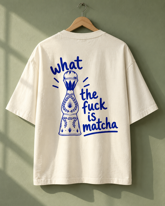An off-white t-shirt hanging on a wooden hanger against a muted sage green wall, featuring a blue graphic of a chess piece and the text 'what the fuck is matcha'.