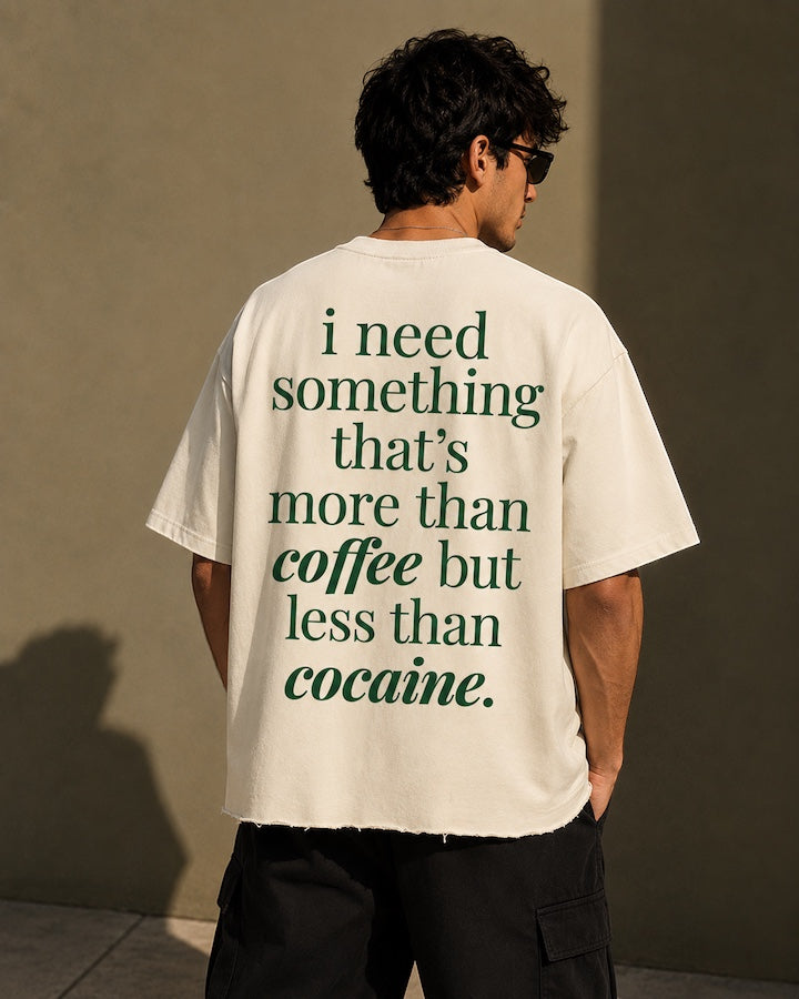 Back view of an off-white boxy fit t-shirt with a raw unfinished bottom hem. It features large green text reading i need something that's more than coffee but less than cocaine.