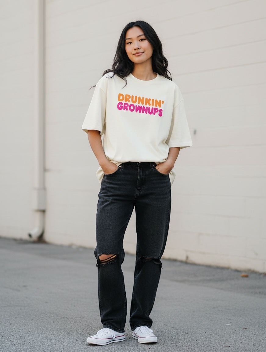 Frolic Saga oversized cream cotton t-shirt with "Drunkin' Grownups" graphic in orange and pink. Model wears black ripped jeans.
