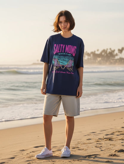 Frolic Saga navy blue oversized cotton tee with retro "Salty Moms Beach Club" graphic and neon pink/teal accents. Model on beach.