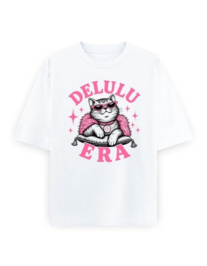 Frolic Saga white 100% cotton oversized t-shirt with a graphic of a cat wearing sunglasses and a pink fluffy boa, sitting on a pillow. Text reads DELULU ERA.