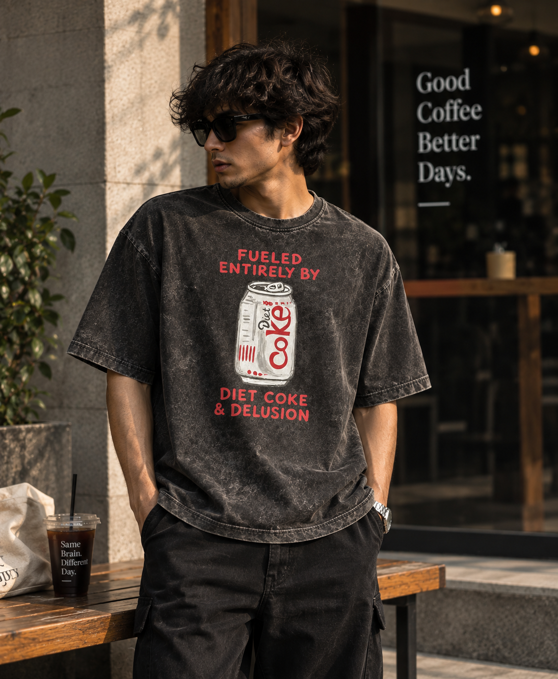 A man wearing a faded black graphic t-shirt with a vintage-style Coke can print and red text, standing outdoors in front of a cafe storefront.